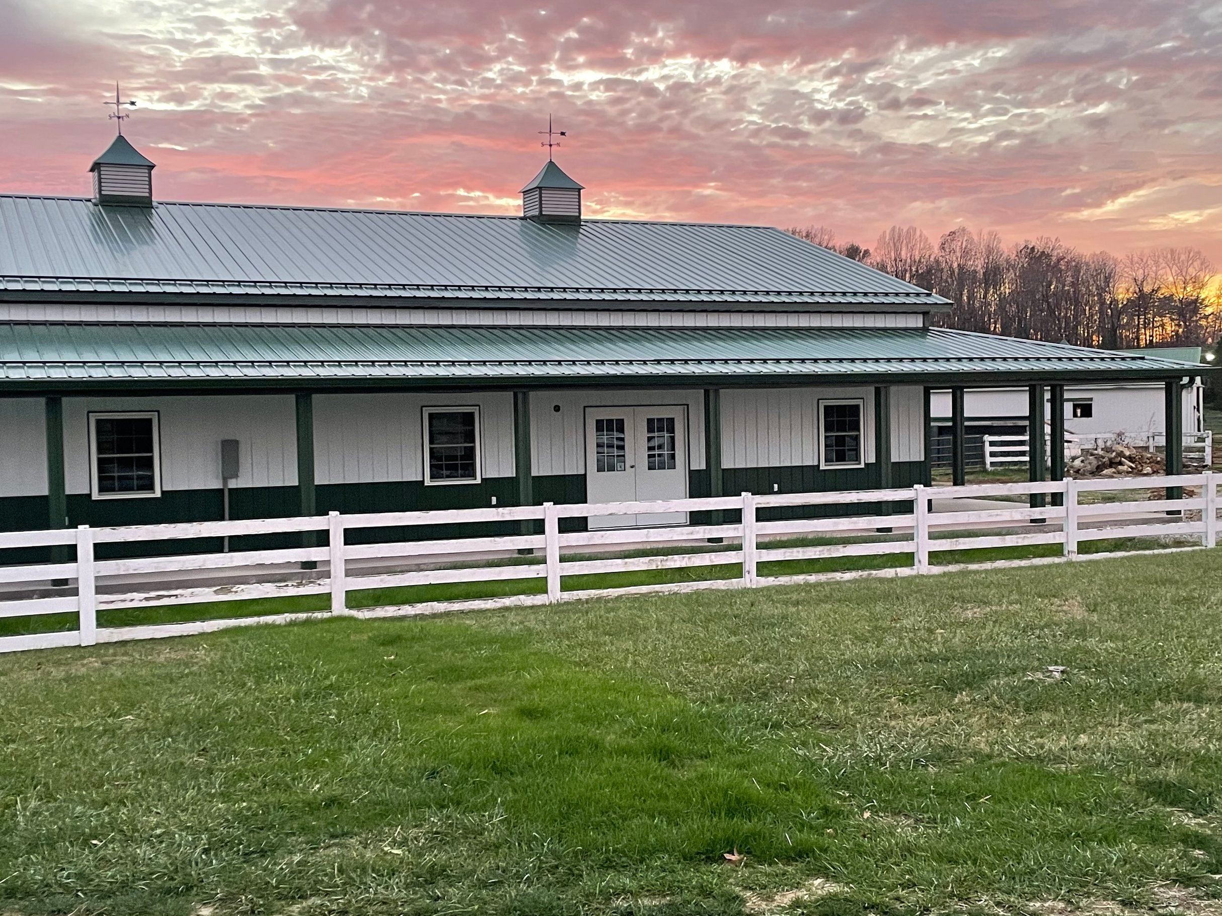Modern horse barn with a green and white exterior, featuring a metal roof and multiple windows, set against a vibrant sunset sky. The barn is surrounded by a white fence and lush green grass, highlighting an inviting equestrian environment.